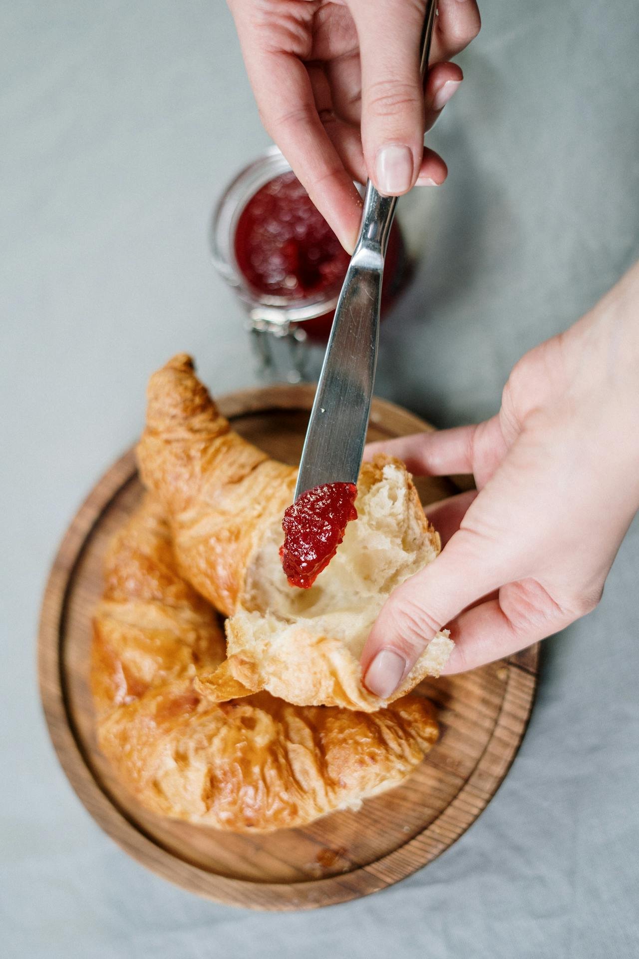 A person enjoys a classic Parisian breakfast, spreading red jam onto a fresh, flaky croissant, illustrating the simple pleasures of French patisserie.